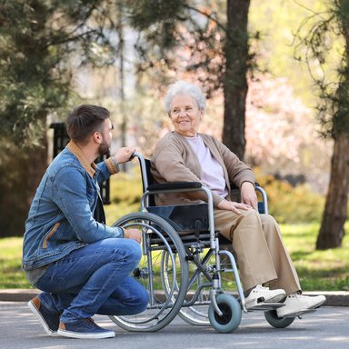 Das Bild zeigt einen jungen Mann bei einer ehrenamtlichen Tätigkeit mit einer Bewohnerin - gemeinsame Rollstuhlfahrt im Park.